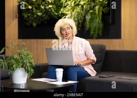 Birassische junge Frau im Büro mit Laptop, während sie auf der Couch sitzt. Arbeiten an einem Projekt in einem modernen Geschäftsbüro mit Pflanzen und Holzakzenten, u Stockfoto