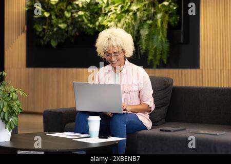 Junge Frau, die an einem Laptop arbeitet, lächelt, während sie im Büro auf der Couch sitzt. Undefinierte Umgebung mit grünem Hintergrund, Pflanzen und AA Stockfoto