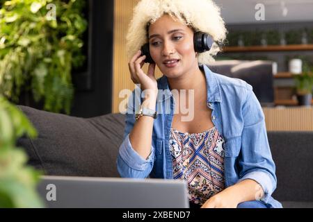 Junge birassische Frau mit Kopfhörern, die im modernen Büro auf den Laptop-Bildschirm schaut. Pflanzen und Holzakzente schaffen einen ruhigen und natürlichen Hintergrund Stockfoto