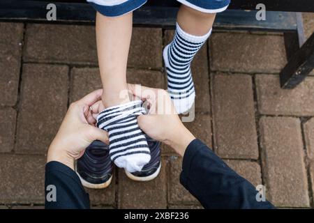 Eine Mutter, die sanft Socken auf ihrem Kleinkind-Sohn in der Bank legt, veranschaulicht das Konzept der Erziehung und Mutterschaft Stockfoto