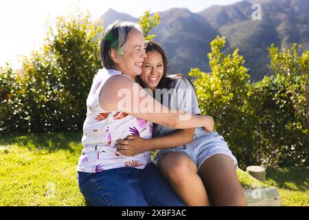 Asiatische Großmutter umarmt birassische junge Enkelin, beide lächeln warm. Sie sitzen draußen mit üppigem Grün und Bergen im Hintergrund, Stockfoto