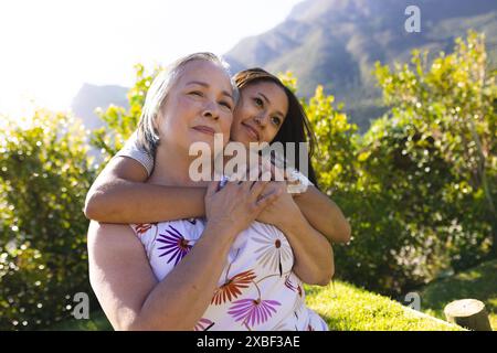 Birassische Enkelin umarmt ihre asiatische Großmutter von hinten, beide lächeln warm Stockfoto