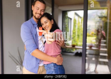 Das junge, vielfältige Paar hält Hausschlüssel und lächelt, kauft das erste Zuhause. Sie stehen vor einem modernen Haus mit großen Fenstern und grüner Stockfoto