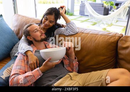 Birassische Frau und kaukasischer Mann entspannen sich auf der Couch und teilen gemeinsam Snacks Stockfoto