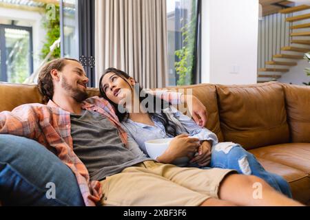 Junge, vielfältige Paare entspannen auf einer Couch. Sie sind in einem modernen Wohnzimmer mit großen Fenstern und Blick auf die Natur, unverändert Stockfoto