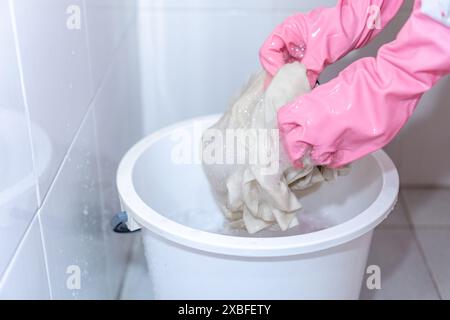 Frau, die Gummihandschuhe trägt, wäscht schmutziges weißes Hemd mit Reinigungsmittel und Aufhellungsmittel in einem Korb Stockfoto