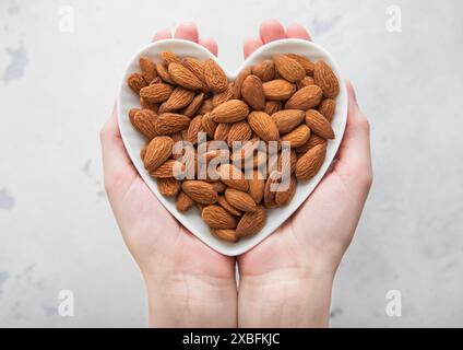 Weibliche Hände, die eine herzförmige Platte mit geschälten Mandelnüssen auf hellem Hintergrund halten. Stockfoto