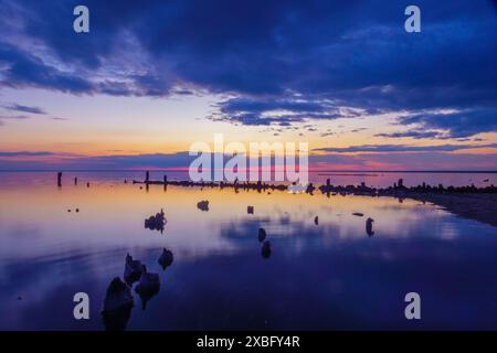 Wunderschöner Sonnenuntergang über einem ruhigen Gewässer, mit dunklen Wolken am Himmel und Resten von Holzpfosten. Friedliche und ruhige Szene. Stockfoto