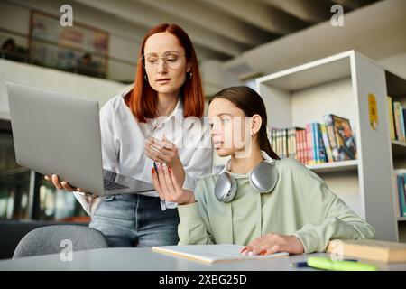 Eine rothaarige Frau unterrichtet ein Teenager-Mädchen in einer Bibliothek, die sowohl engagiert als auch konzentriert auf einen Laptop-Bildschirm für moderne Bildung ist. Stockfoto