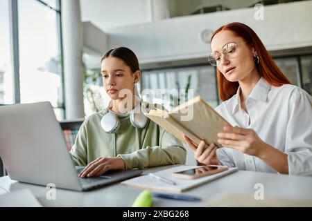 Eine rothaarige Frau unterrichtet ein Teenager-Mädchen an einem Tisch, beide tauchen in einen Laptop ein, während sie ein Buch liest. Stockfoto