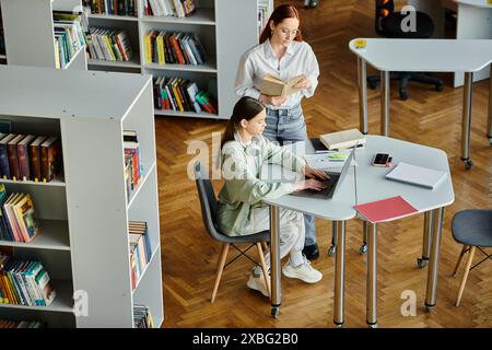 Eine rothaarige Frau unterrichtet ein Teenager-Mädchen, das in einer ruhigen Bibliothek mit einem Laptop an einer Nachschulstunde teilnimmt. Stockfoto