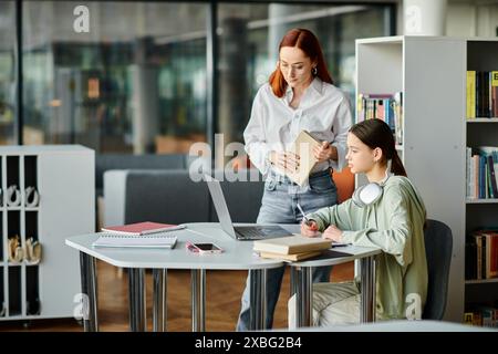 Eine rothaarige Frau unterrichtet ein Mädchen im Teenageralter in einer Bibliothek und arbeitet an einem Laptop für den Unterricht nach der Schule zusammen. Stockfoto