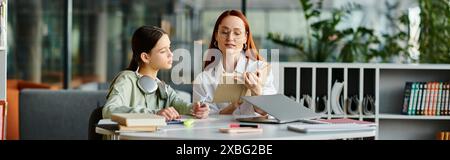 Eine rothaarige Frau unterrichtet ihre Tochter im Teenageralter, die sich beide während des Unterrichts nach der Schule in einem modernen Büro auf einen Laptop konzentrieren. Stockfoto