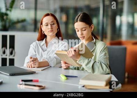 Eine rothaarige Frau unterrichtet ein Teenager-Mädchen an einem Tisch, beide in ein Buch eingetaucht, Laptop in der Nähe. Moderne Bildung in Aktion. Stockfoto