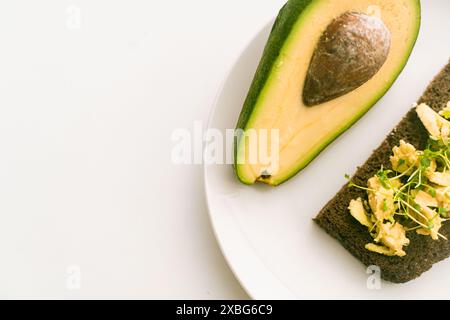 Eine Scheibe oder ein Toast von Roggenbrot auf einem Teller mit Guacomol mit Mikrogrün und einer halben Avocado auf einer weißen Platte auf hellem Hintergrund. Gesund, diätetisch Stockfoto