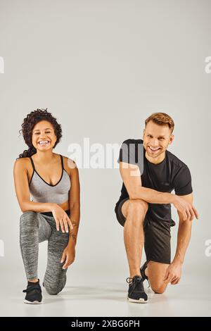 Junge interrassische Sportpaare, die sich in aktiver Kleidung auf grauem Hintergrund in einem Studio-Setting zusammenhocken. Stockfoto