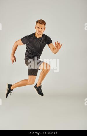 Ein junger athletischer Mann in einem schwarzen T-Shirt springt energisch in einem Studio mit grauem Hintergrund. Stockfoto
