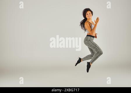 Eine junge Afroamerikanerin in athletischer Kleidung springt freudig vor grauem Hintergrund in einem Studio-Setting. Stockfoto