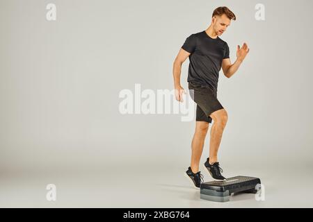 Ein junger Sportler in aktiver Kleidung benutzt einen Stepper in einem Studio vor grauem Hintergrund. Stockfoto