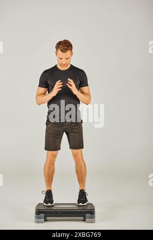 Ein junger Sportler in aktiver Kleidung fordert sein Gleichgewicht auf einem Stepper in einem Studio mit grauem Hintergrund heraus. Stockfoto