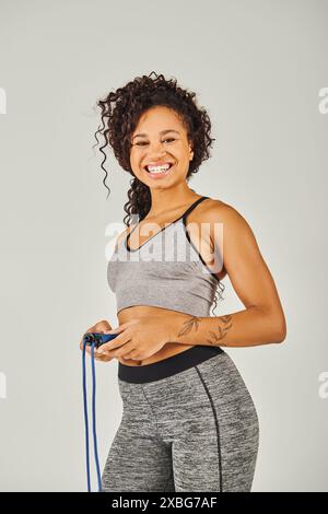 Eine lockige afroamerikanische Sportlerin in Activewear lächelt, während sie in einem Studio vor grauem Hintergrund ein Seil hält. Stockfoto