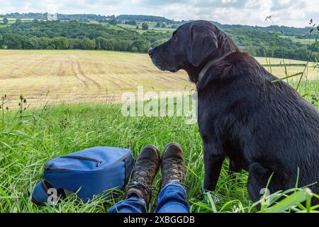 Ein schwarzer labrador-Retriever, der neben seinem Besitzer am Rande eines Feldes sitzt. Die Wanderschuhe des Besitzers und ein Rucksack befinden sich neben dem Hund. Stockfoto