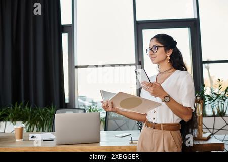 Junge indianerin in Brille steht am Schreibtisch und hält Mappe. Stockfoto