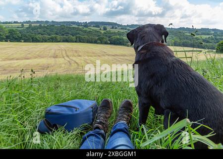 Ein schwarzer labrador-Retriever, der neben seinem Besitzer am Rande eines Feldes sitzt. Die Wanderschuhe des Besitzers und ein Rucksack befinden sich neben dem Hund. Stockfoto