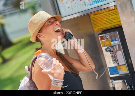 Eine fröhliche Frau, die am Telefon an einem Münztelefon im Freien telefoniert, während sie in Miami eine Flasche Wasser hält Stockfoto