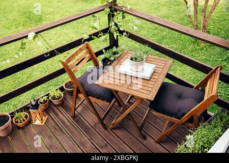 Kleiner Balkon mit Holzbrettern und Gartenmöbeln für zwei Personen, Holzstühlen und Tisch. Verschiedene Kräuter, die in Töpfen wachsen: basilikum, Zwiebeln, Rosmarin. Stockfoto