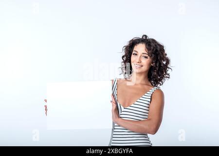 Schöne lächelnde mediterrane Frau in ihren 20, die ein Schild mit Platz für Werbetexte auf weißem Hintergrund hält. Das Bild hat viel Kopierraum Stockfoto