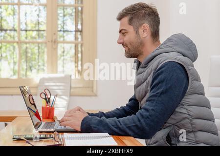 Profilporträt eines Mannes mittleren Alters, der an seinem Laptop arbeitet, während er an seinem Schreibtisch in einem hellen Büro sitzt. Er ist konzentriert und engagiert in seine Aufgaben, Highl Stockfoto