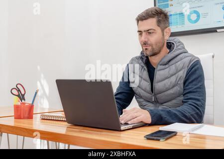 Ein attraktiver reifer Mann mit einem rasierten Bart, der an seinem Schreibtisch sitzt, arbeitet konzentriert und blickt aufmerksam auf den Bildschirm. Stockfoto