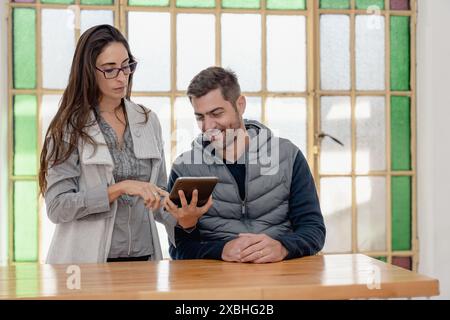 Zwei Kollegen diskutieren, während sie in einem hellen Büro ein Tablet verwenden. Der Mann sitzt an einem Holztisch und die Frau, die neben ihm steht und einen pflegt Stockfoto