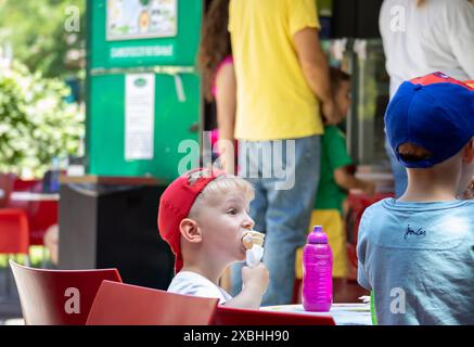 Kleine Kinder genießen an einem sonnigen Tag Eis in einem Café im Freien, umgeben von farbenfrohen Hintergründen und Menschen Stockfoto