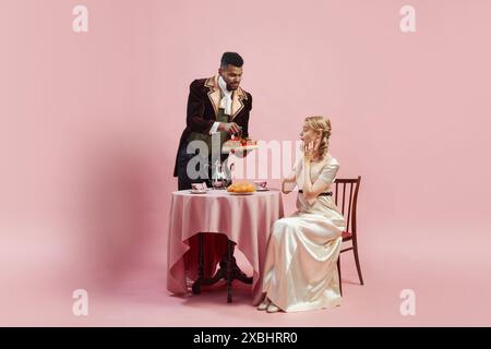 Mann in reicher Samtjacke bietet Tablett mit frischen Erdbeeren für elegante Frau in Satinkleid vor rosa Studiohintergrund Stockfoto