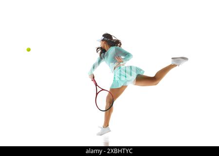 Sportliche Frau in türkisfarbenem Tenniskleid springt vorwärts, um den Ball zu treffen, während der Action vor weißem Studiohintergrund festgehalten wird. Stockfoto