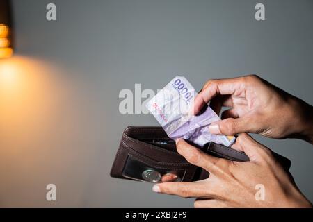 Eine Person, die eine Banknote der indonesischen Rupiah 10.000 und eine braune Brieftasche hält. Der Hintergrund ist verschwommen, und links befindet sich eine warme Lichtquelle. Stockfoto