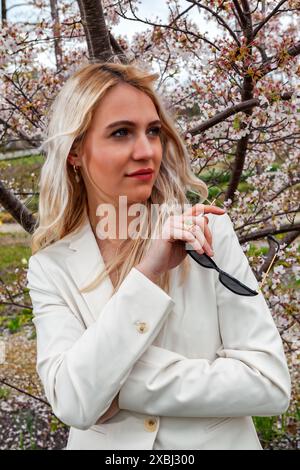 Ein Porträt einer jungen, blonden Frau, die eine Sonnenbrille hält. Im Hintergrund ist ein blühender Sträucher, der eine frühlingshafte und lebhafte Stimmung erzeugt. Die Stockfoto