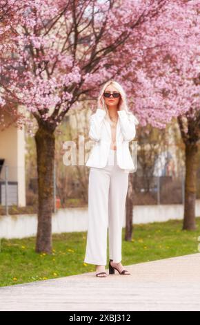 Eine stilvolle Frau im weißen Hosenanzug steht vor einem blühenden Baum und zieht ihre Sonnenbrille an. Die Szene verbindet natürliche Schönheit mit Fashionik Stockfoto