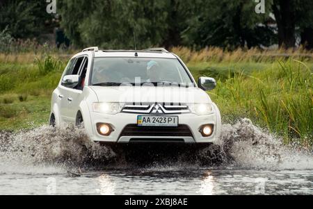 Weißer Geländewagen, der einen Fluss durchquert. Spritzer streuen in alle Richtungen. Mitsubishi Pajero Sport überquert einen flachen Teich, der von Scirpus-Pflanzen umgeben ist. Stockfoto