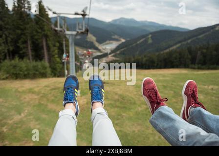 Aus Sicht: Fahrt im Sessellift in Bergsteigen. Beine eines Paares, das in einem Sessellift fährt. Wandern mit Sessellift. Stockfoto