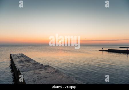 Küste mit wenigen Piers und Silhouetten von Fischern bei Sonnenaufgang. Ruhiges Wasser, Möwen ruhen auf dem Betonpier, Sonnenaufgang über dem Meer. Stockfoto