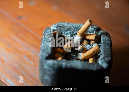 Eine Nahaufnahme eines schwarzen Aschenbechers, gefüllt mit Zigarettenkippen auf einem Holztisch. Stockfoto