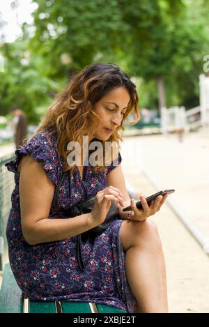 Eine hübsche hispanische Frau, die ihr Handy benutzt, während sie in einem Park in Paris sitzt. Stockfoto