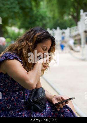 Ein Nahporträt einer hübschen hispanischen Frau, die gähnt, während sie ihr Handy in einem Park in Paris ansieht. Kopierbereich Stockfoto