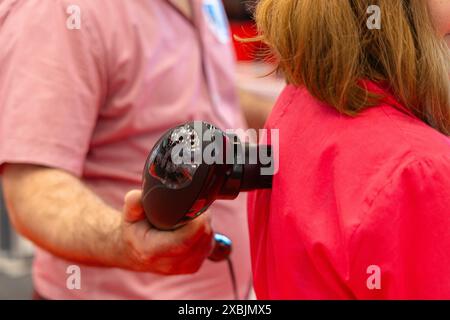 Osijek Kroatien - 7 06 2024: Mann in einem rosa Hemd demonstriert auf einer Messe die Verwendung einer Massagepistole bei Frauen. Die Frau trägt einen roten Shi Stockfoto