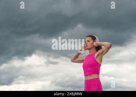 Sportliche Frau, die Musik mit Kopfhörern hört, pinkfarbene Sportkleidung vor einem dramatischen Himmel trägt Stockfoto