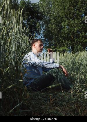Porträt eines Mannes in einem grünen ukrainischen Feld: Vertikaler Full-Length Profile Face Angle Shot eines jungen Mannes in einem Sky Blue Shirt und Green Jeans sitzend Stockfoto