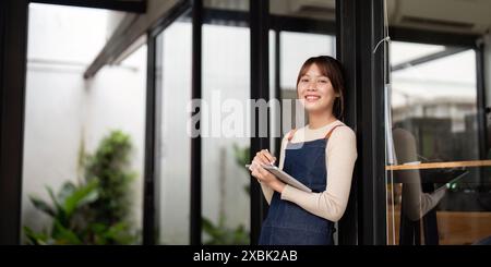 Junge Frau, die ein kleines Geschäft als Cafe-Besitzer beginnt, lächelnd und mit Notizbuch in der Hand, steht im modernen Cafe-Eingang, bereit, Kunden zu begrüßen Stockfoto